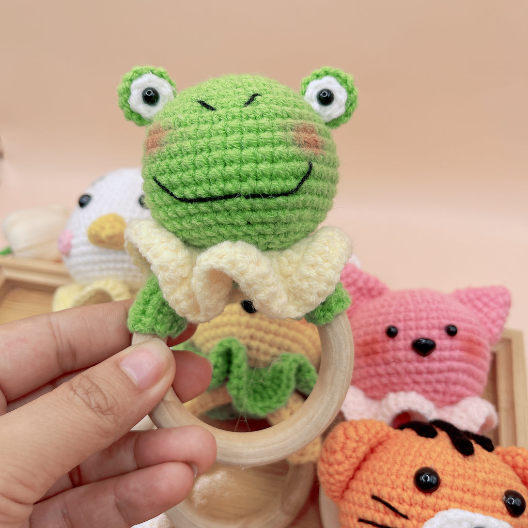 Hand holding green frog Animal Crochet Rattle with wooden ring and other crochet animals in background