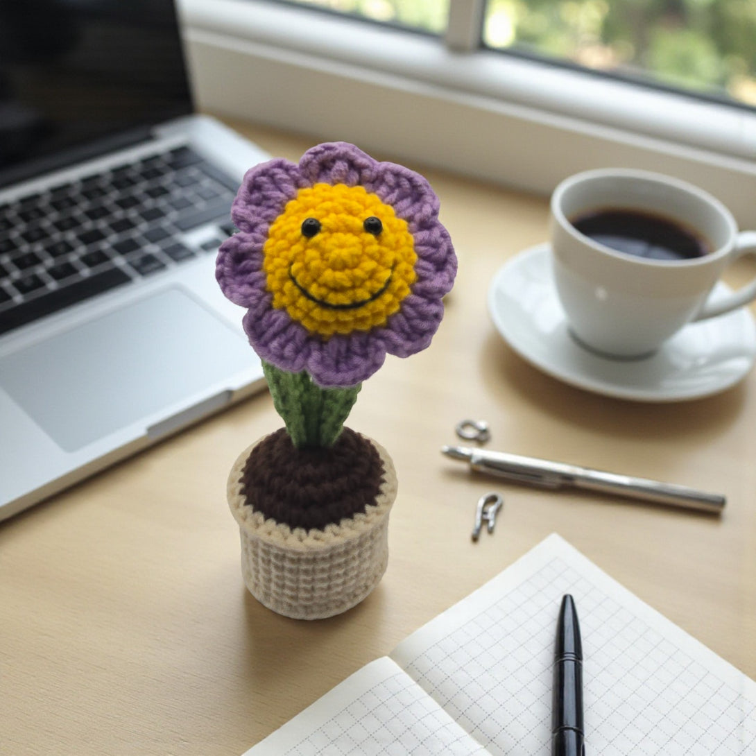 Colorful crochet flower pot featuring a smiling flower in purple and yellow on a desk with coffee