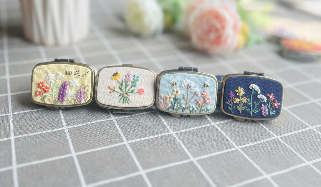 Stitchery Zone embroidered jewelry box collection with floral designs and pastel colors on a gray grid surface