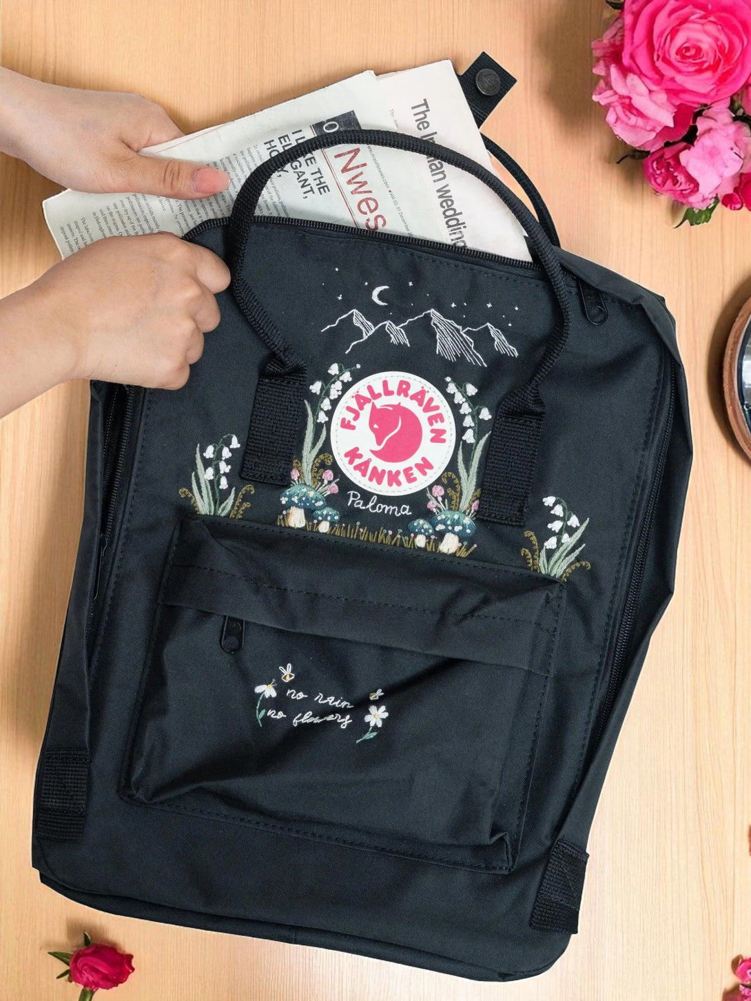 Stitchery Zone black Fjallraven Kanken Classic Backpack with embroidered mountains and flowers hands placing newspaper