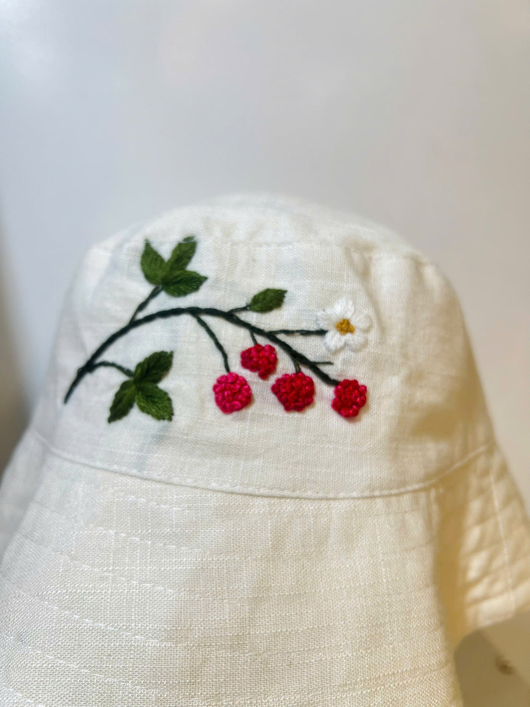 Hand Embroidered Berry Linen Baby Hat by Stitchery Zone with red berries and green leaves on white linen fabric