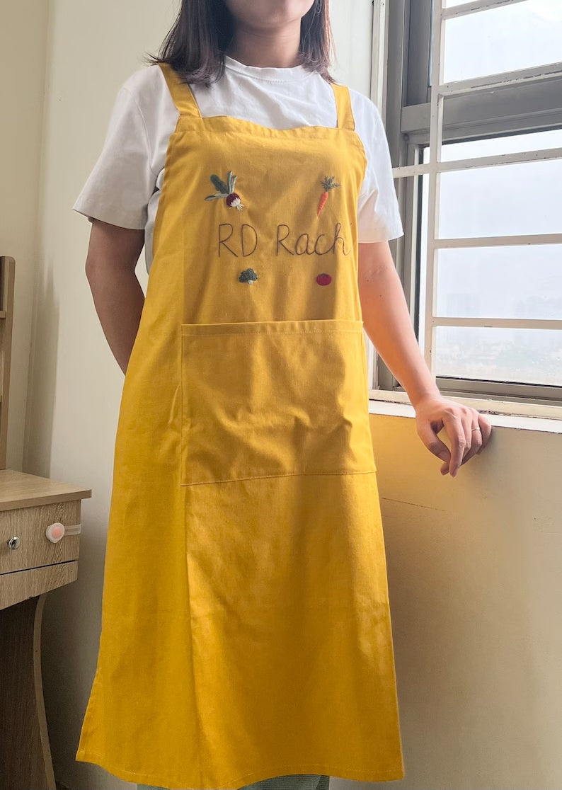 Stitchery Zone yellow Vegetable Embroidery Linen Apron worn by person standing near window
